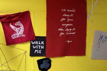 Red tote bags with white Indigenous art and a Walk With Mi banner hanging in front of a yellow wall